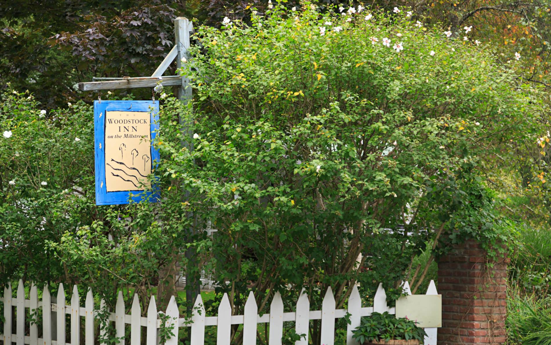 Beige and blue Woodstock Inn sign hanging from rustic wood post over a white picket fence surrounded by greenery