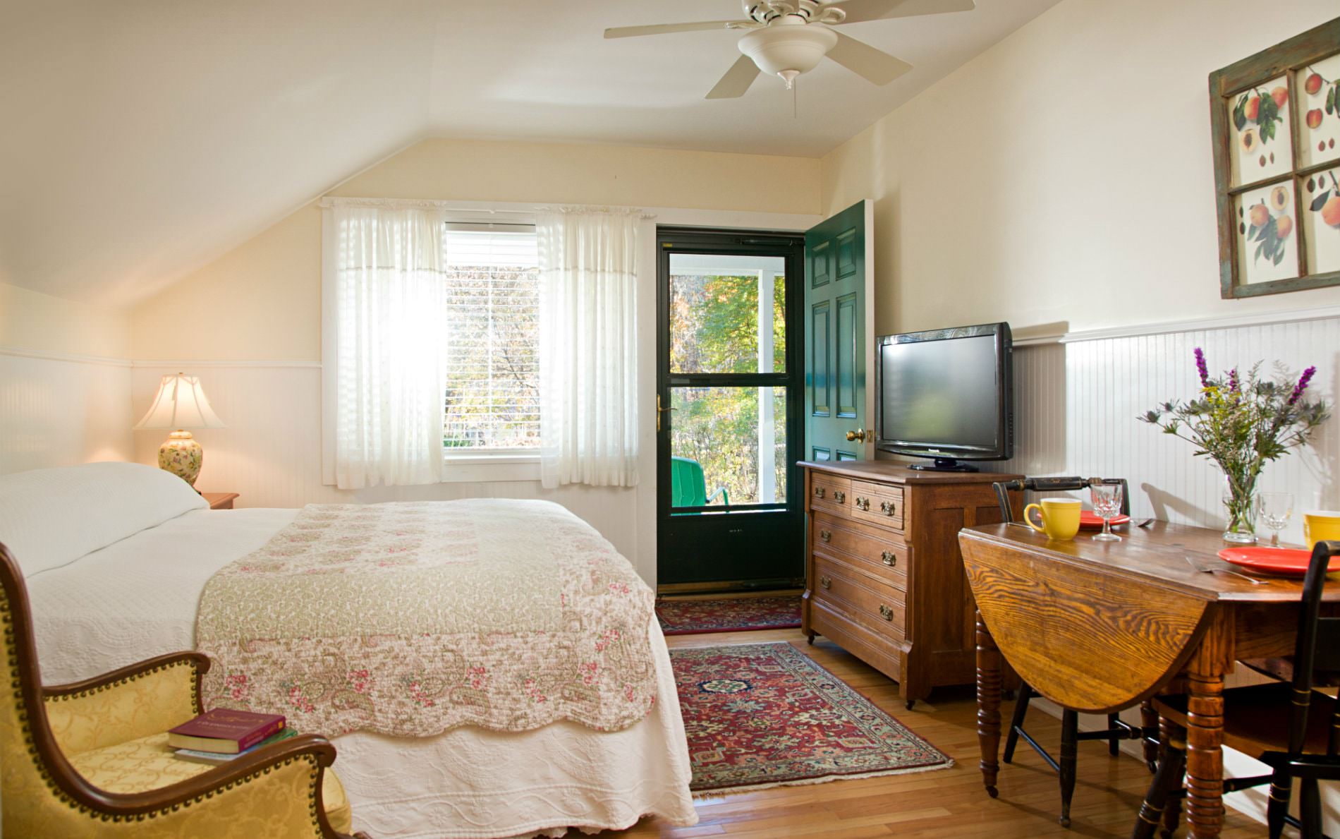 Beige room with wood floor, glass door, window, white covered bed, side chair, drop-leaf table and dresser with tv