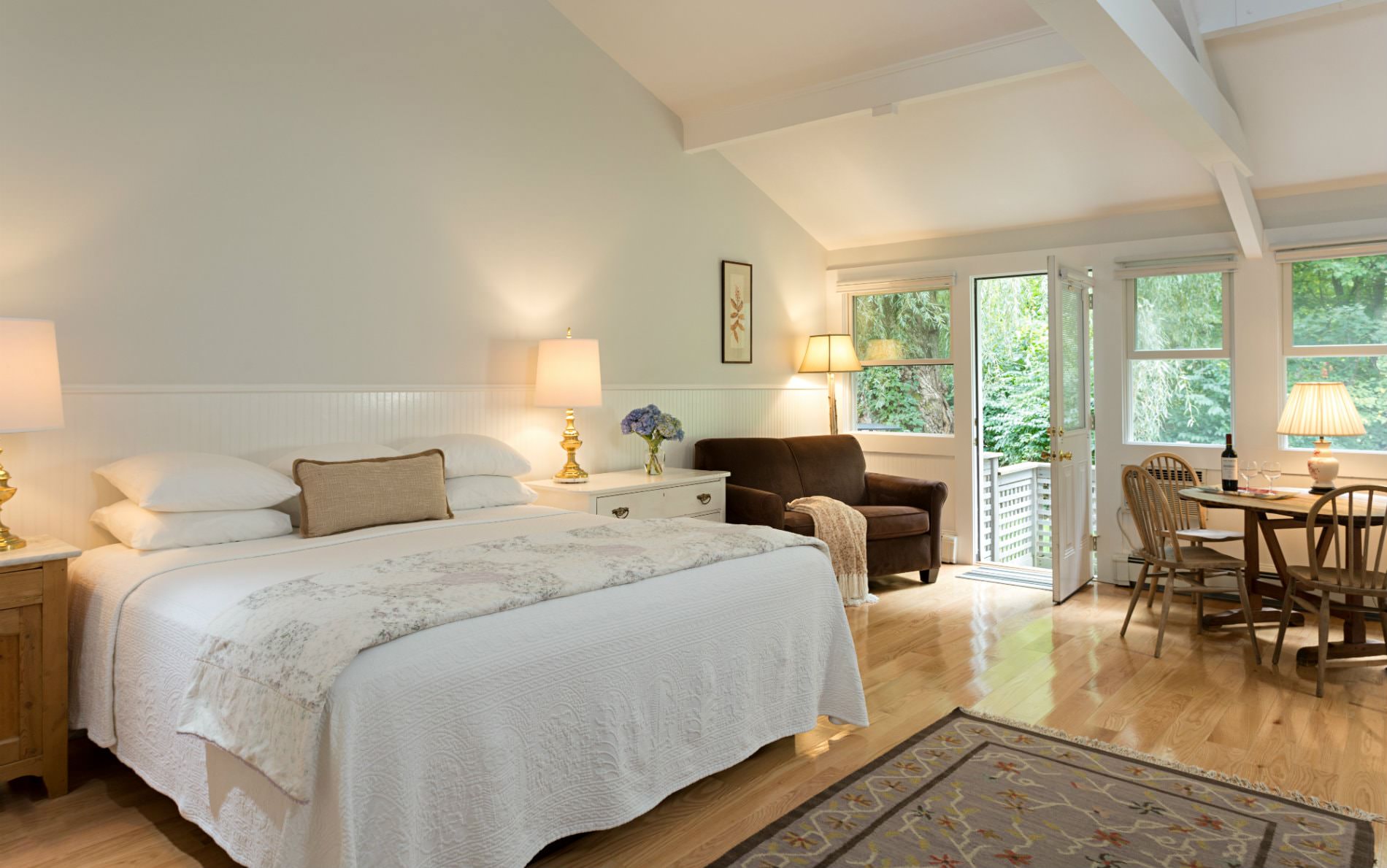 White vaulted room with wood floors, several windows, glass door, white covered bed, love seat, and table and chairs