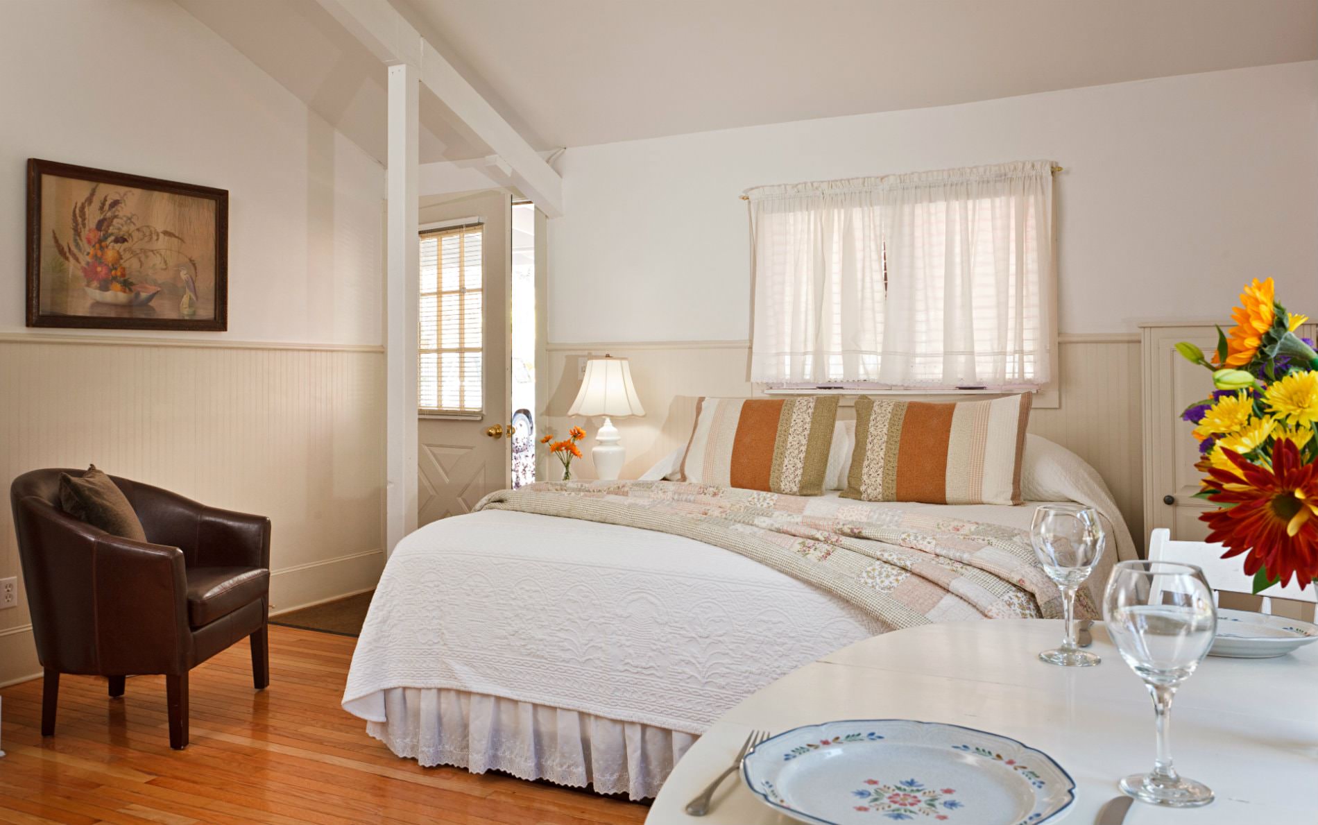 Beige vaulted room with wood floors, white covered bed, leather club chair, and white table with fresh flowers