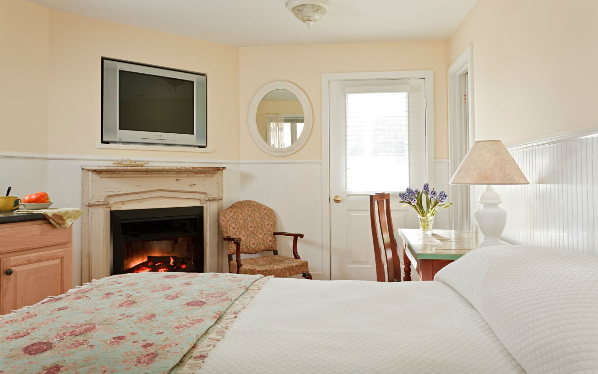 Beige and white room with glass door, flat screen tv, fireplace, side chair, desk and chair and white covered bed