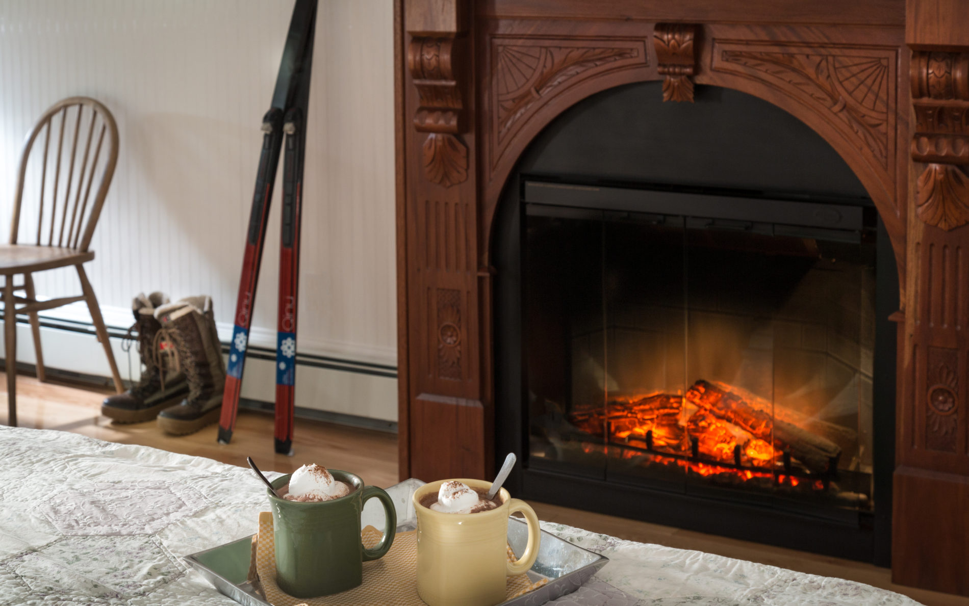 a tray on a bed with two mugs of hot cocoa, a fireplace in teh background with orange flames and a pair cross country skis leaning against the white wall.