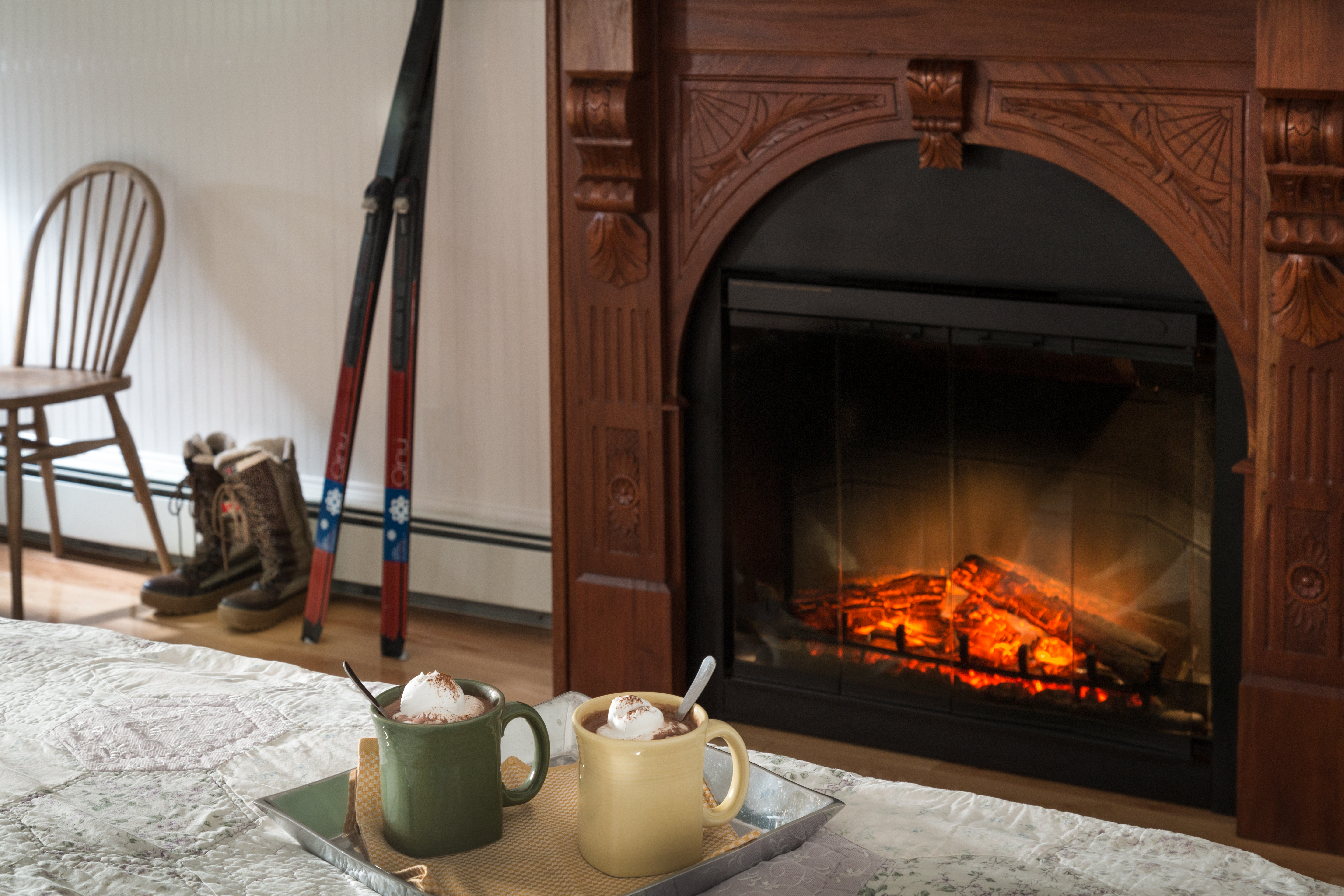 a tray on a bed with two mugs of hot cocoa, a fireplace in teh background with orange flames and a pair cross country skis leaning against the white wall.