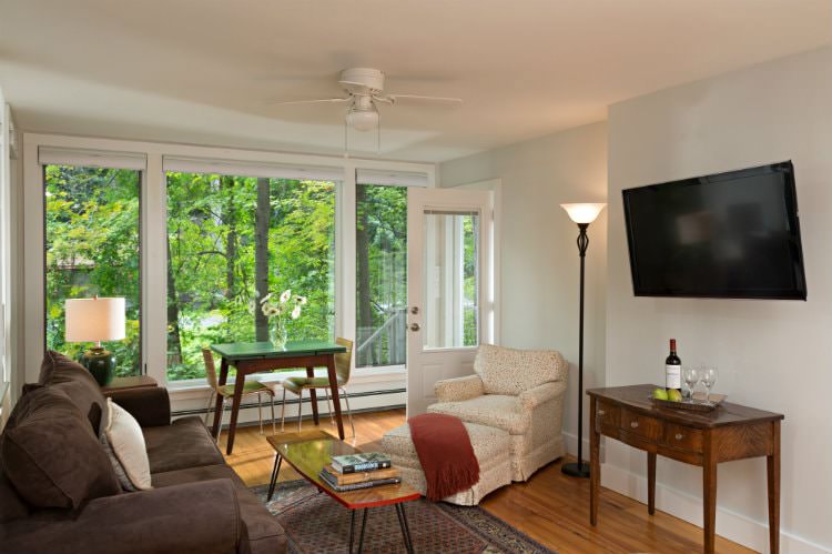 White room with wood floors, large triple window, brown sofa and white upholstered chair with ottoman around flat screen tv