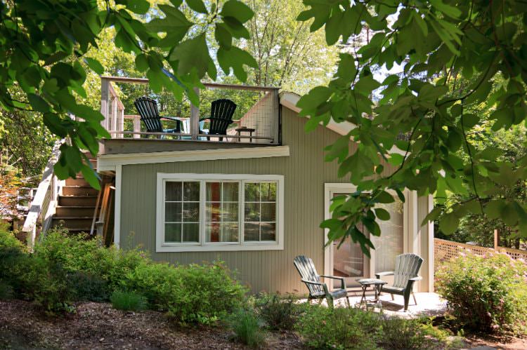 Tan and white cottage, small patio with two chairs, rooftop deck with chairs, surrounded by green trees