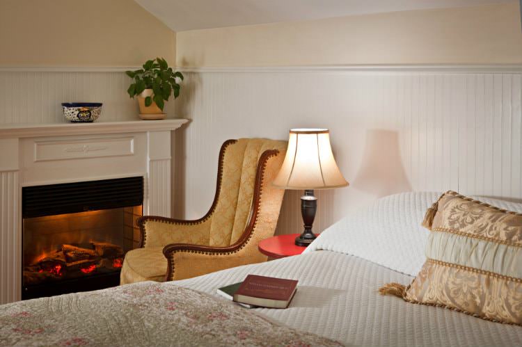 Beige room with white bead board, white covered bed, upholstered side chair in front of a fireplace