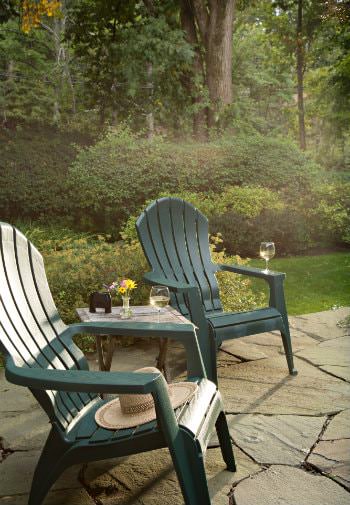 Flagstone patio with two green Adirondack chairs and table surrounded by green grass, shrubs and trees