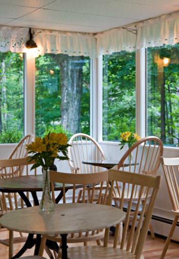 Wood tables and chairs topped with fresh yellow flowers surrounded by windows overlooking lush greenery