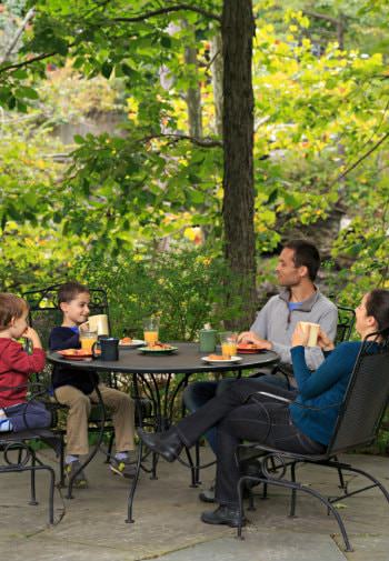 Father, mother, and two small boys sitting outside at a black metal patio table and chairs surrounded by green trees