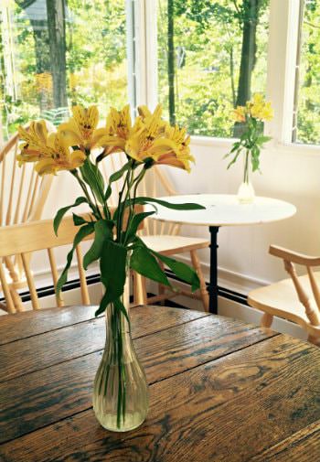 Wooden tables and chairs topped with glass vases of yellow flowers surrounded by windows overlooking green trees