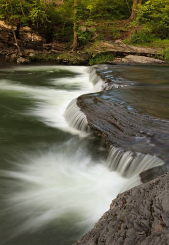 Shallow waterfall with white spraying water surrounded by woods and green leafed trees