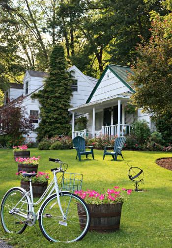 Adirondack chairs, round tubs of pink flowers, and white bicycle on green lawn - tall trees and white cottages in background