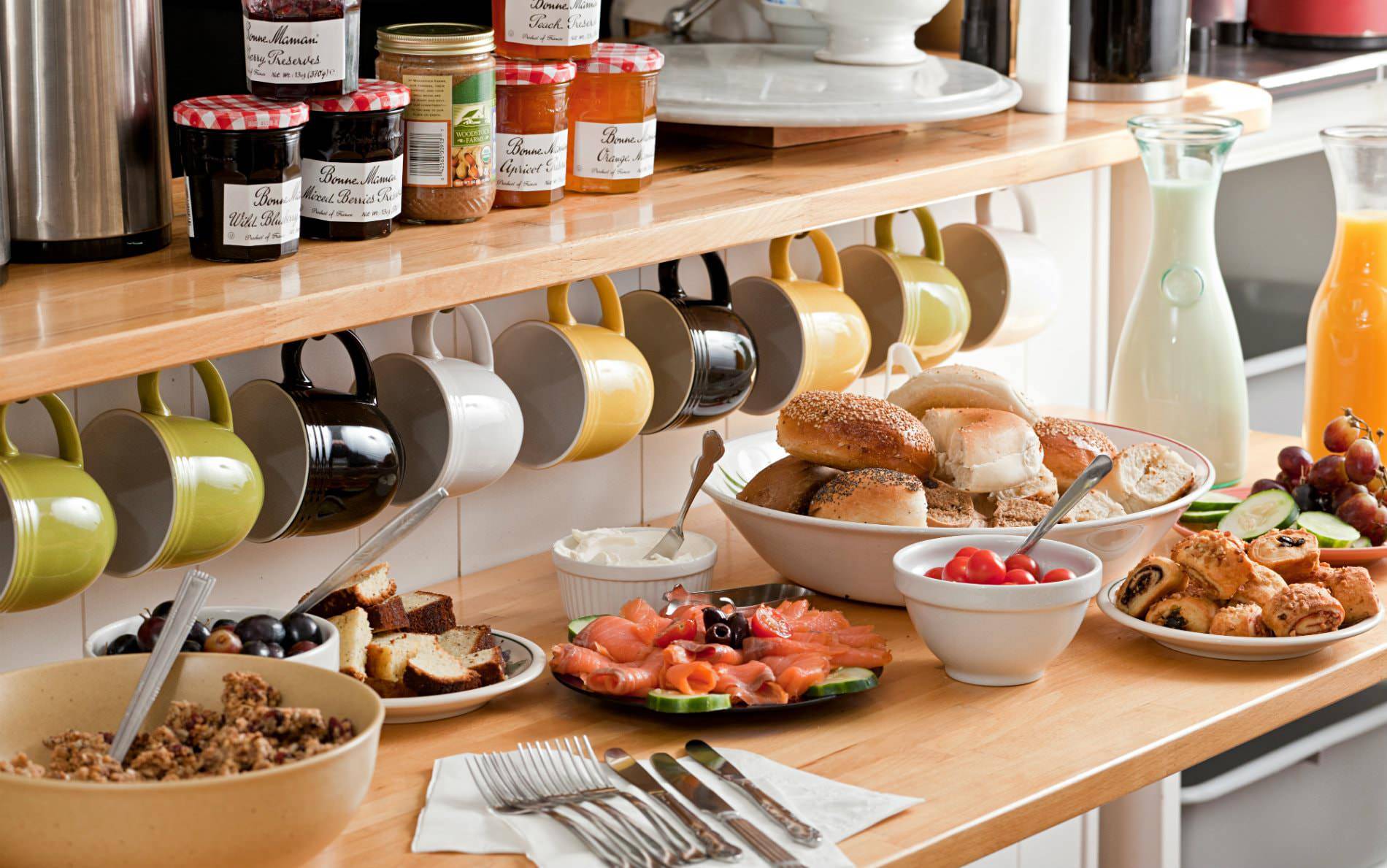 Butcher block counter topped with breakfast foods under a row of colorful mugs hanging from a wood shelf