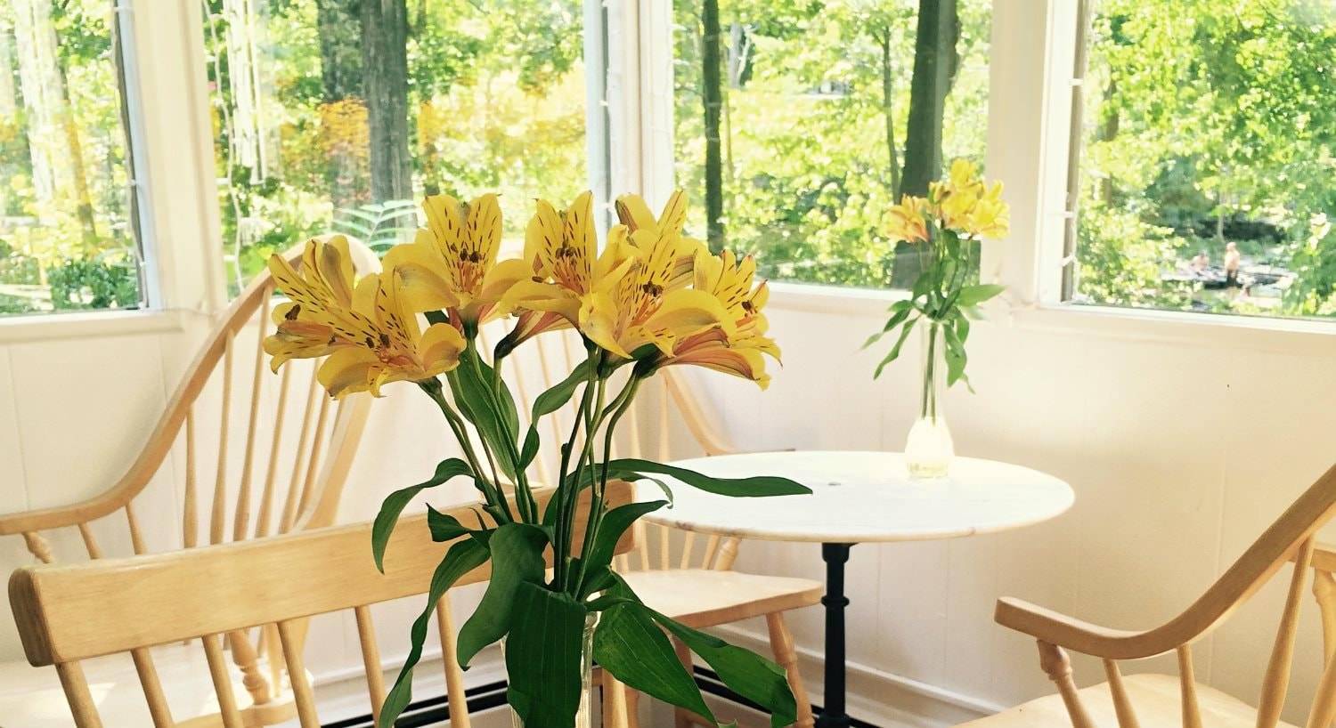 Small wood dining tables topped with vases of yellow lilies surrounded by windows with view of lush greenery