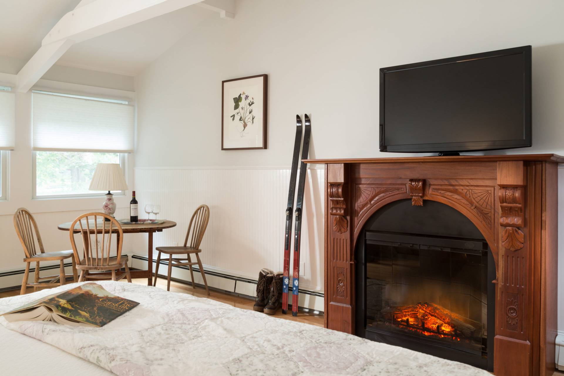 Fire burning in wooden fireplace, skis and boots lean on white wall with a book on a whote bedspread and a wooden table with wine bottles. 