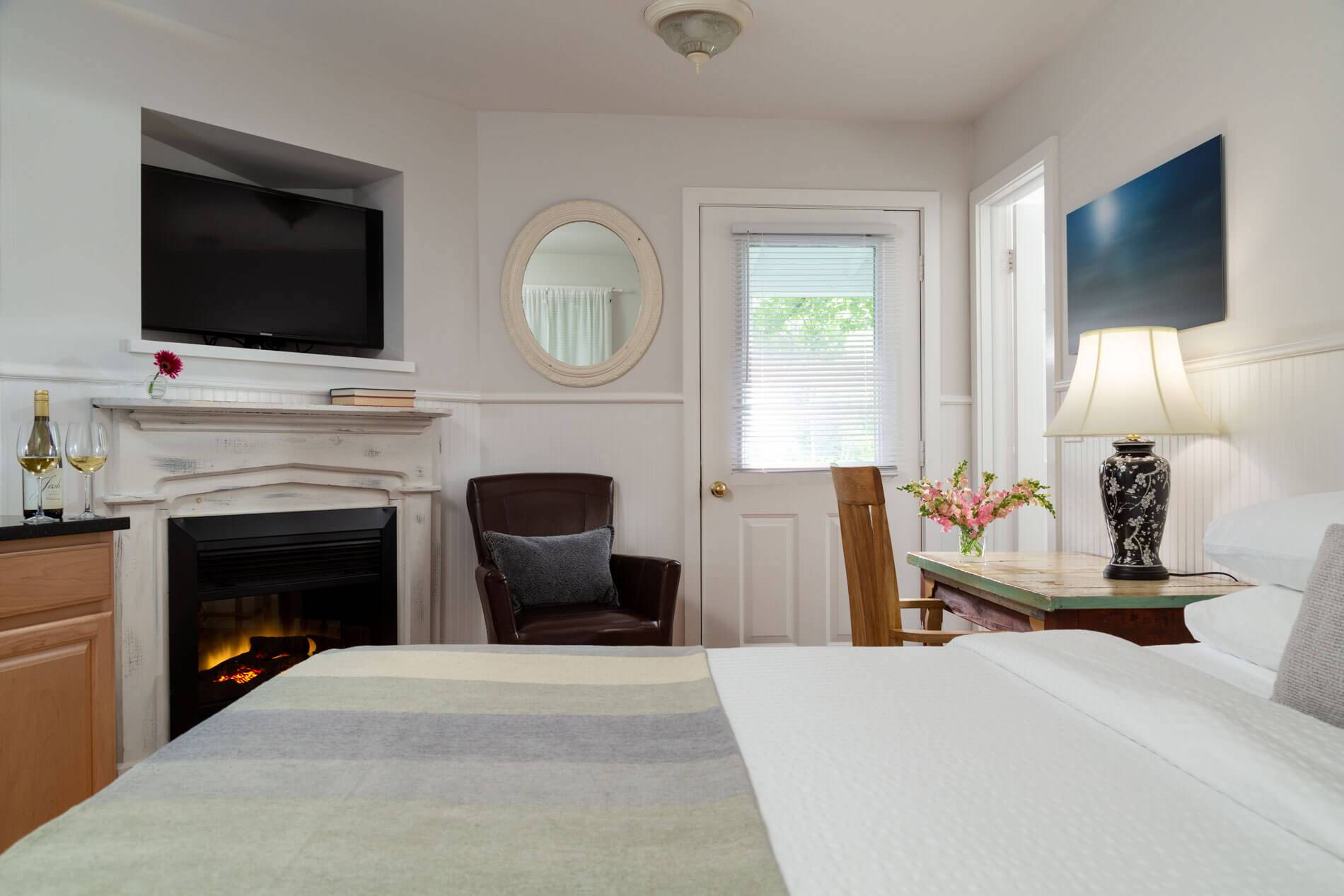 A bed with a white cover and green blanket at the foot is in the foreground, then in the room there's an electric fireplace , a black chair and a door with a sun filled window